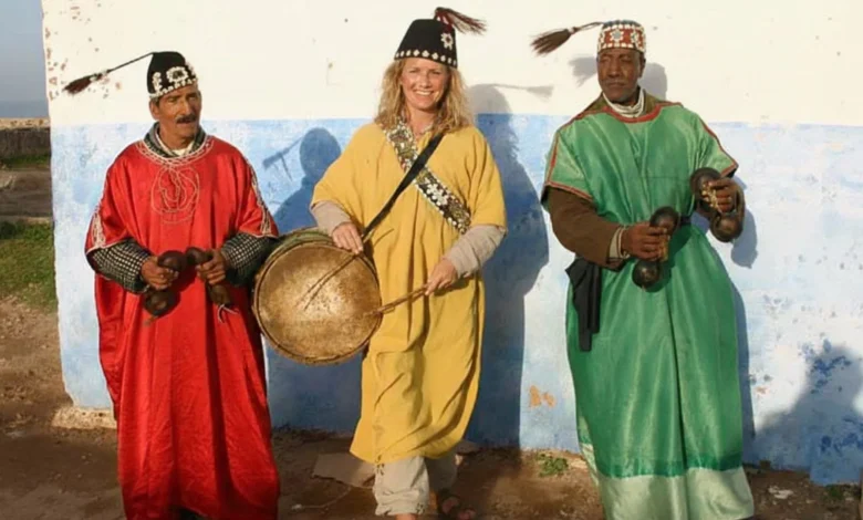 Smiling solo female traveller wearing traditional yellow Gnawa attire and holding a Guembri instrument, standing between two Moroccan musicians in red and green costumes holding Qraqeb castanets - a scene embodying Moroccan hospitality and proving is Morocco safe for solo female travellers - authentic photo enhanced with AI