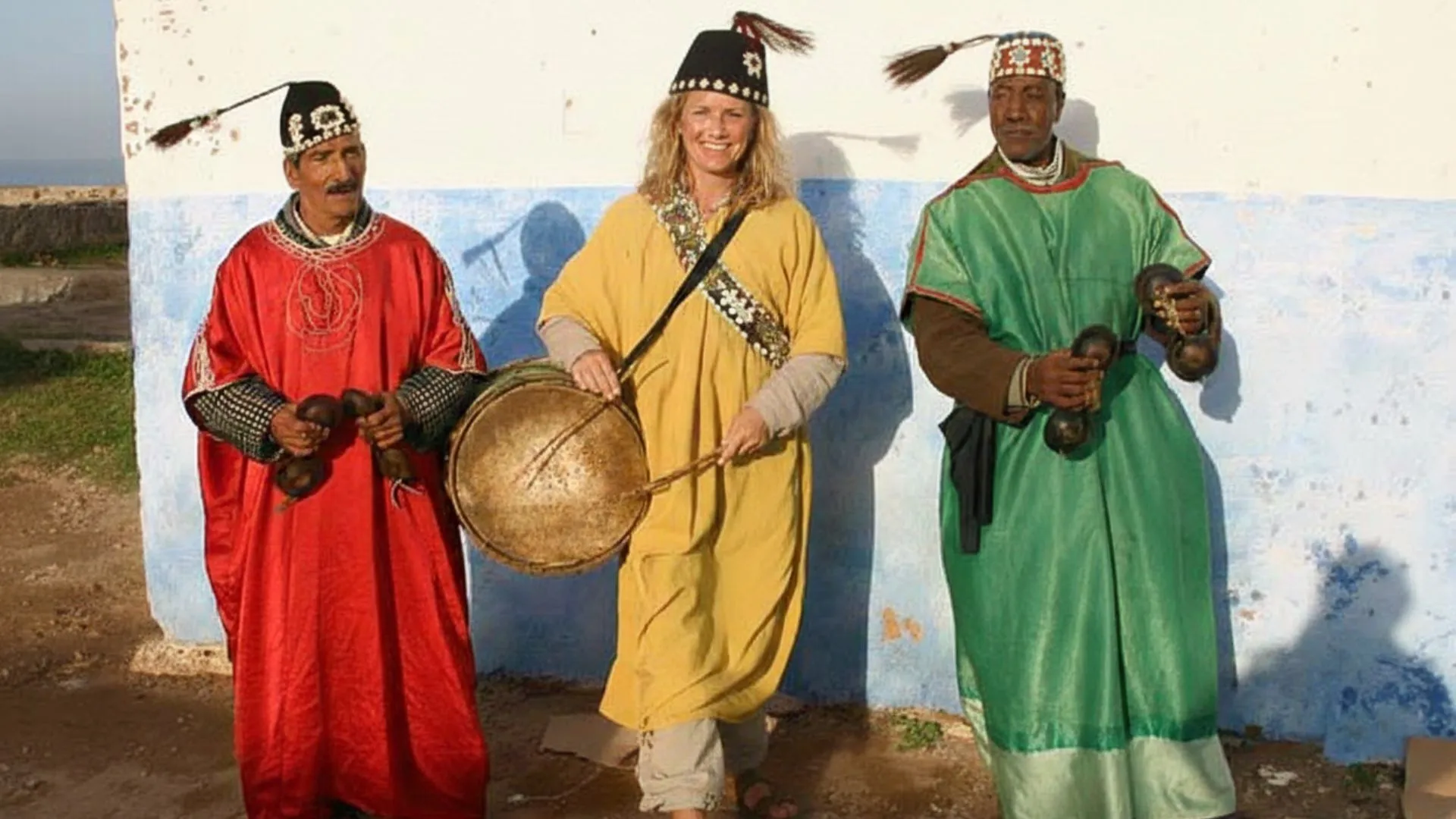 Smiling solo female traveller wearing traditional yellow Gnawa attire and holding a Guembri instrument, standing between two Moroccan musicians in red and green costumes holding Qraqeb castanets - a scene embodying Moroccan hospitality and proving is Morocco safe for solo female travellers - authentic photo enhanced with AI