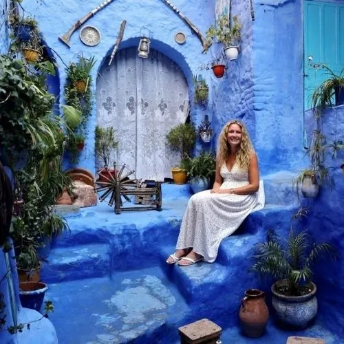 Solo female traveller wearing a long white dress sits on blue-painted steps in Chefchaouen's famous alleys - proving is Morocco safe for solo female travellers with this peaceful scene surrounded by blue walls, traditional Moroccan door, and green potted plants  - authentic photo enhanced with AI
