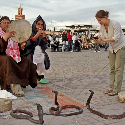 Solo female traveller taking photos of snake charmers at the famous Jemaa el-Fnaa square in Marrakech, with traditional Moroccan musicians, cobras, and Koutoubia Mosque minaret in the background - a scene reflecting is Morocco safe for solo female travellers - authentic photo enhanced with AI