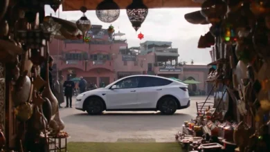 A white Tesla Model Y positioned in a traditional Moroccan marketplace alley, flanked by ornate metal lanterns and hanging handicrafts, captured from Tesla's official Morocco launch promotional video