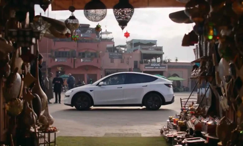 A white Tesla Model Y positioned in a traditional Moroccan marketplace alley, flanked by ornate metal lanterns and hanging handicrafts, captured from Tesla's official Morocco launch promotional video
