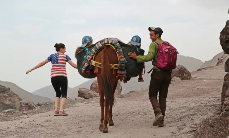 American tourist wearing a US flag shirt hiking with a Moroccan guide and a mule in the Atlas Mountains, illustrating that Morocco is safe for Americans and welcoming to adventurous travelers.
