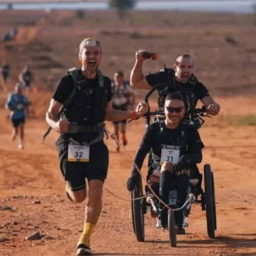 MDS Handi team finishing the Marathon des Sables 2026 with a wheelchair athlete and two assistants celebrating on a desert trail in Morocco