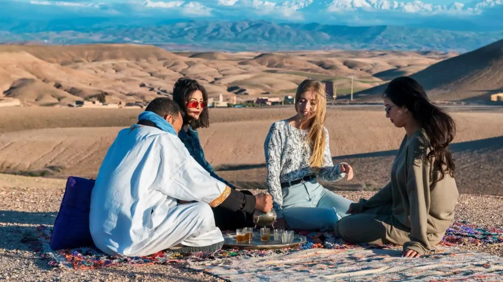 Amazigh host pouring traditional mint tea for travelers seated on a colorful Berber rug in the Moroccan desert with Atlas Mountains in the background