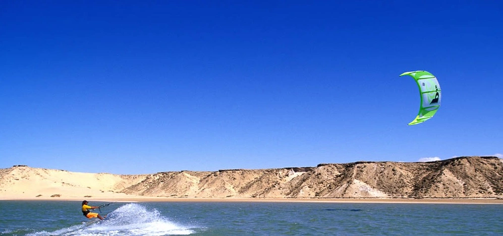 Kitesurfer gliding across the flat lagoon in Dakhla Morocco with golden sand dunes under a clear blue sky
