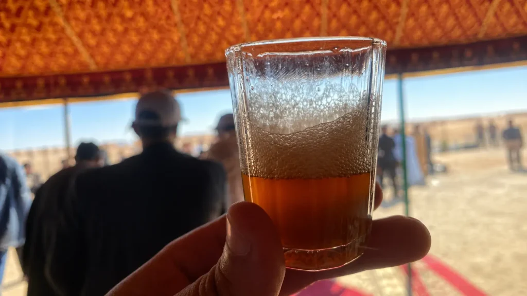 A glass of Moroccan mint tea with traditional foam crown held under a desert tent in southern Morocco