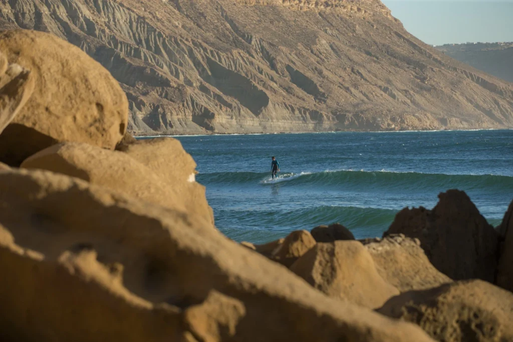 Surfer riding a wave at Taghazout Morocco with rocky cliffs and mountain backdrop along the Atlantic coast
